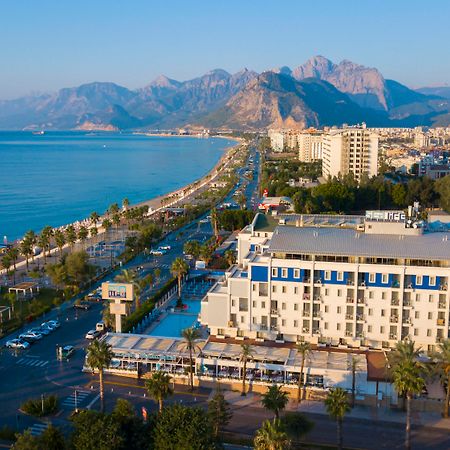 Sealife Family Resort Hotel Antalya Exterior photo The photo depicts a scenic coastal view, featuring a beachfront area lined with palm trees. In the foreground, there is a hotel or resort with a pool and outdoor seating. The road runs parallel to the beach, with some vehicles visible. In the backgro