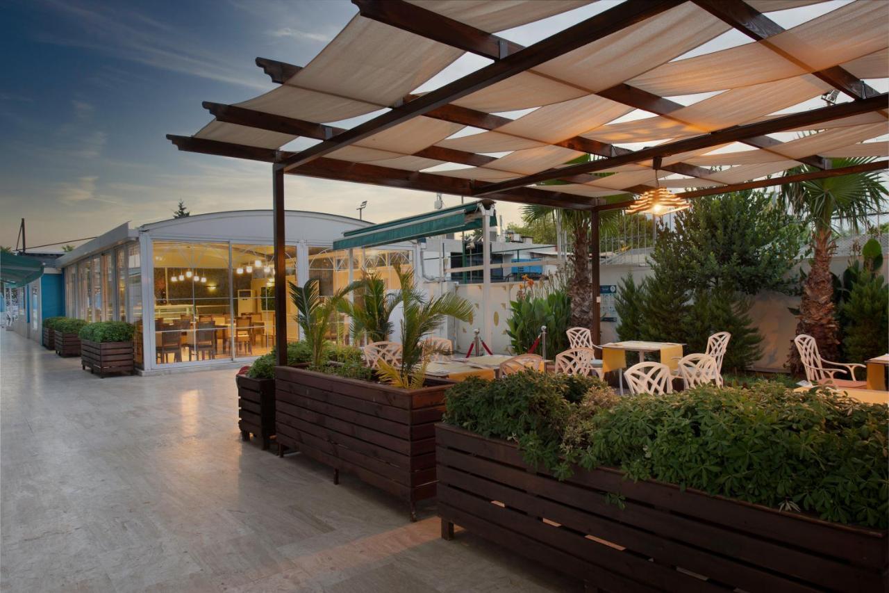 Sealife Family Resort Hotel Antalya Exterior photo The photo depicts an outdoor space, likely a café or restaurant terrace. It features a wooden structure with a grid-like canopy that provides shade, and several white chairs with decorative designs are arranged around small tables. There are large pl