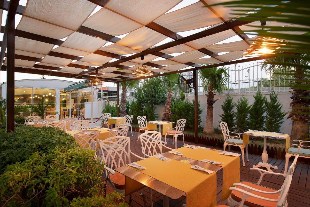 Sealife Family Resort Hotel Antalya Exterior photo The photo depicts an outdoor dining area of a restaurant featuring a pergola with fabric coverings that provide shade. The space is arranged with several tables adorned with bright yellow tablecloths. The chairs are designed with a distinctive white 