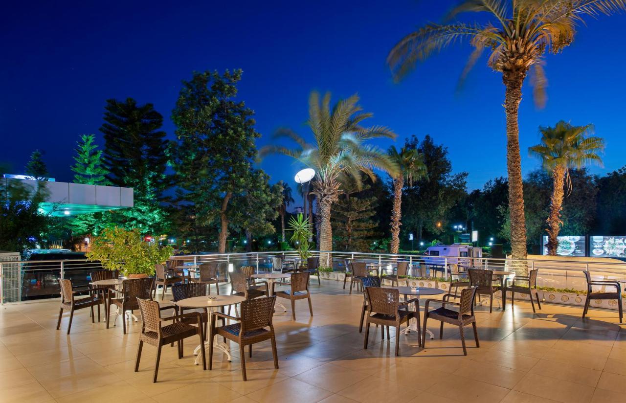 Sealife Family Resort Hotel Antalya Exterior photo The photo depicts an outdoor dining area, likely part of a restaurant or café, set in the evening. There are several tables with chairs arranged across a tiled patio. The space is surrounded by tall palm trees and greenery, creating a tropical ambian