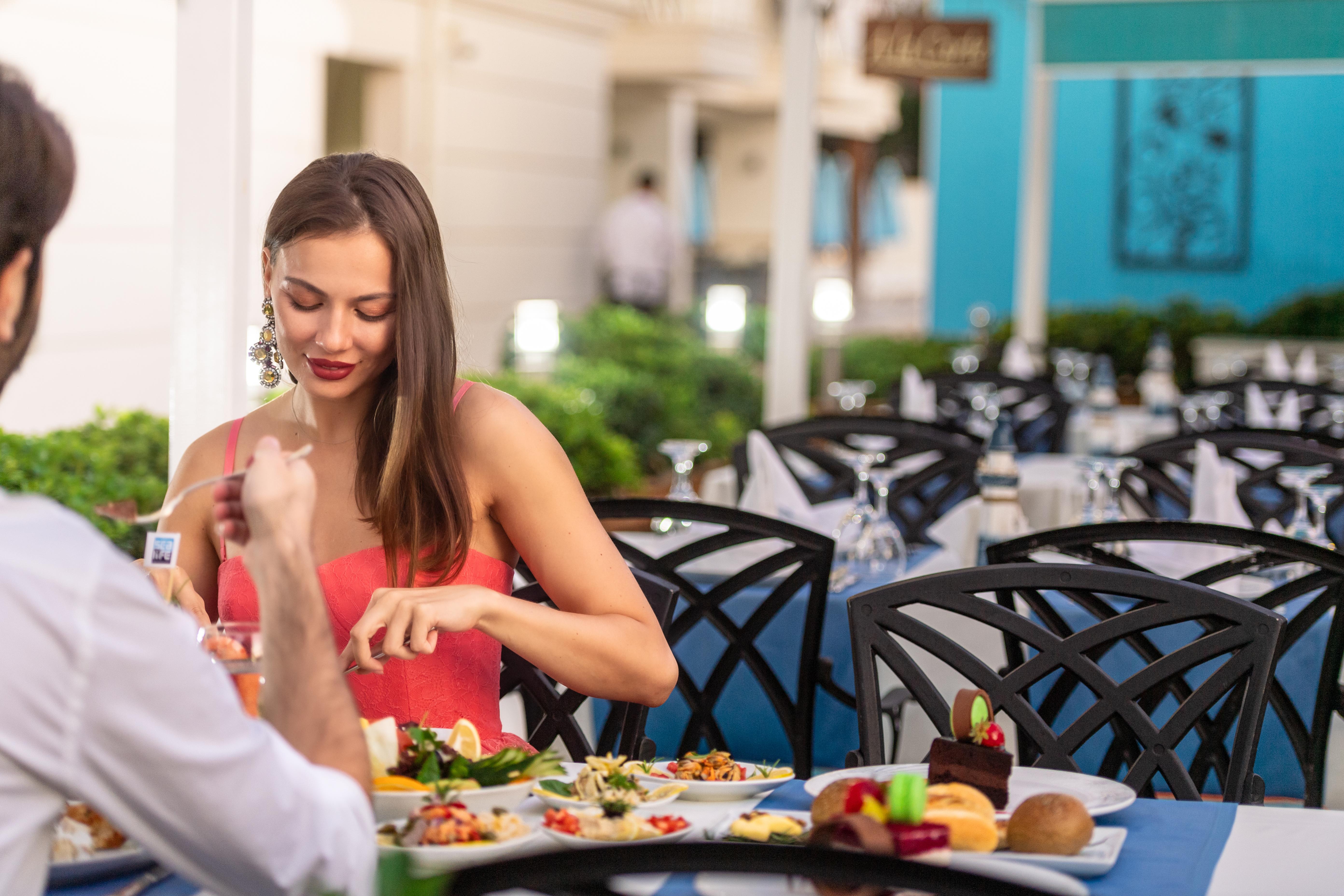 Sealife Family Resort Hotel Antalya Exterior photo The photo shows a dining scene at a restaurant. A woman in a red dress is seated at a table, interacting with her meal. She appears to be taking time to enjoy her food. The table is set with various dishes, including salads and desserts. The backgrou