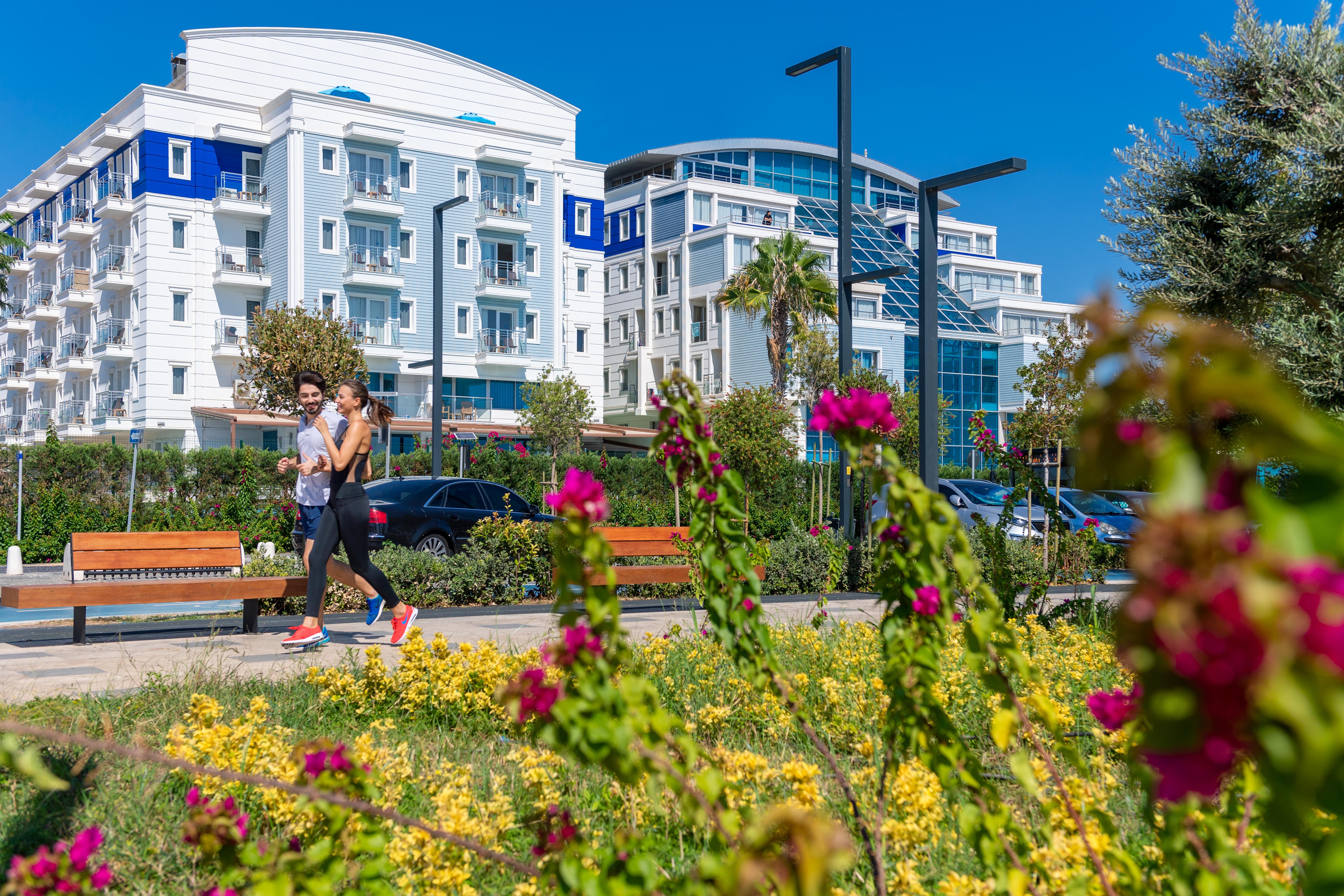 Sealife Family Resort Hotel Antalya Exterior photo The photo depicts a sunny outdoor scene featuring two individuals walking along a path. They are surrounded by a vibrant landscape filled with yellow flowers and pink bougainvillea. In the background, there are modern buildings with balconies, some h