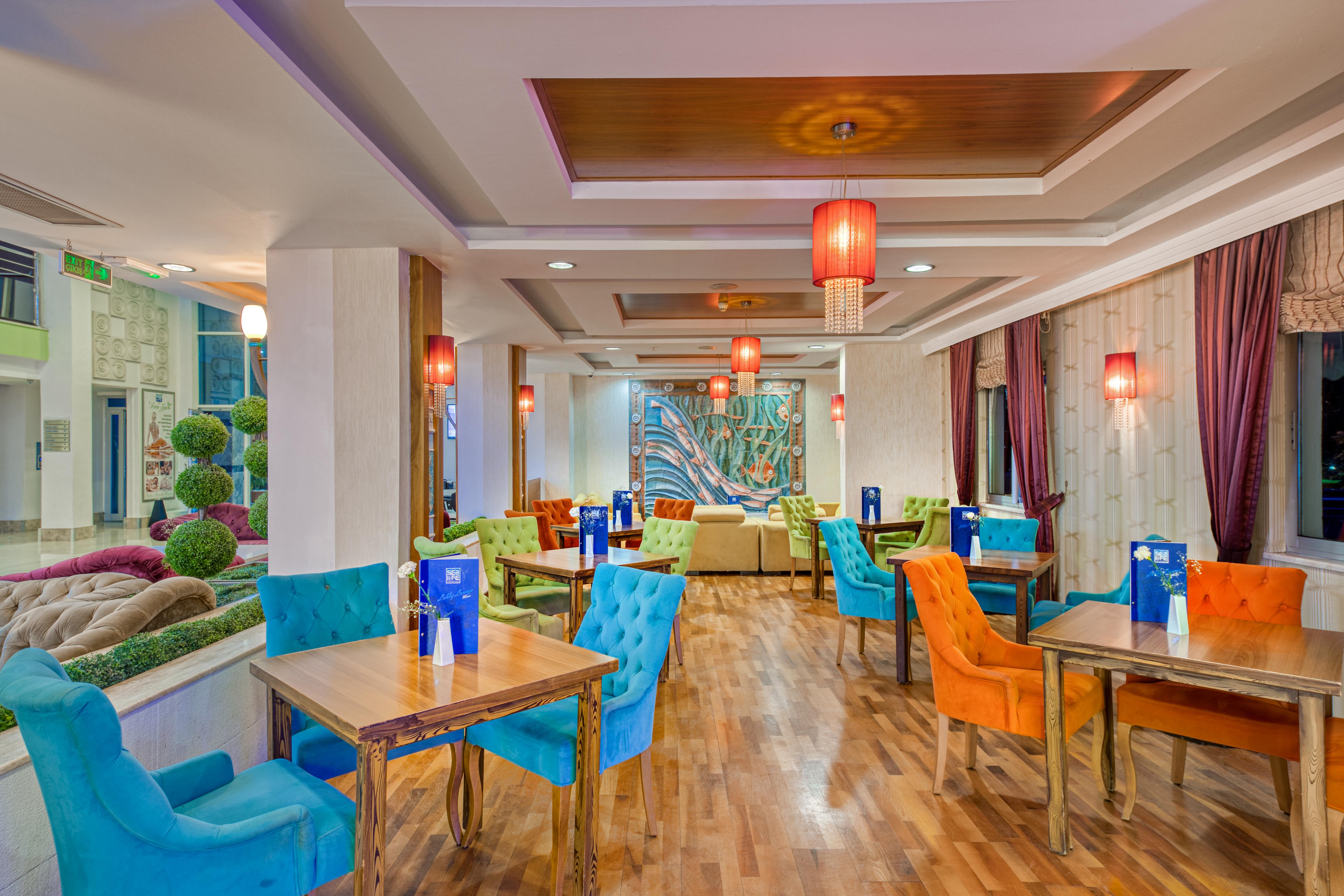 Sealife Family Resort Hotel Antalya Exterior photo The photo shows a stylish dining area with a contemporary design. The space features a combination of colorful chairs in blue and orange, and wooden tables arranged throughout. The walls have a warm, neutral tone and are complemented by decorative el