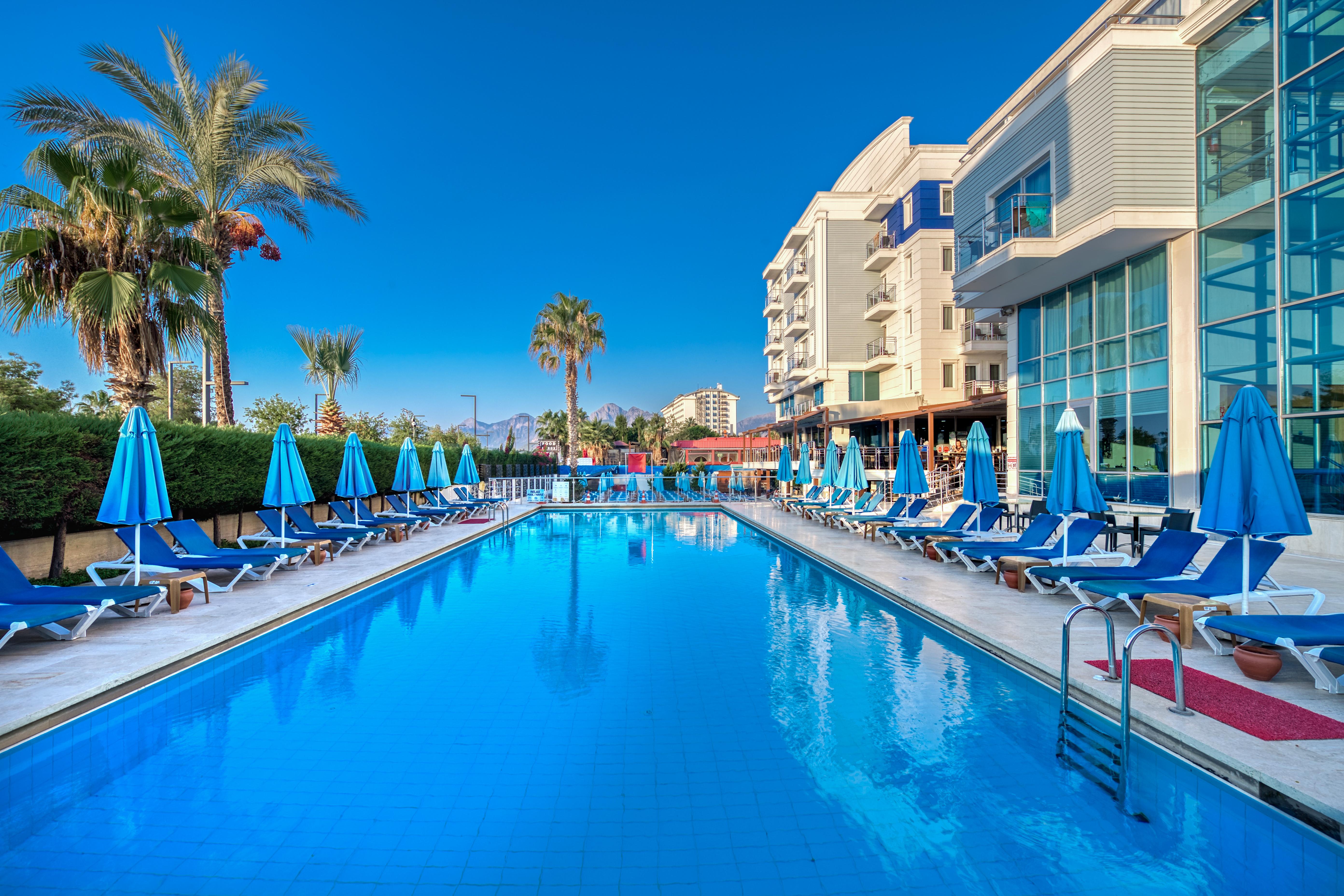 Sealife Family Resort Hotel Antalya Exterior photo The photo depicts a serene poolside scene at a hotel or resort. In the foreground, a large blue swimming pool reflects the clear sky, with comfortable lounge chairs arranged neatly around it under blue umbrellas. Lush palm trees add to the tropical a