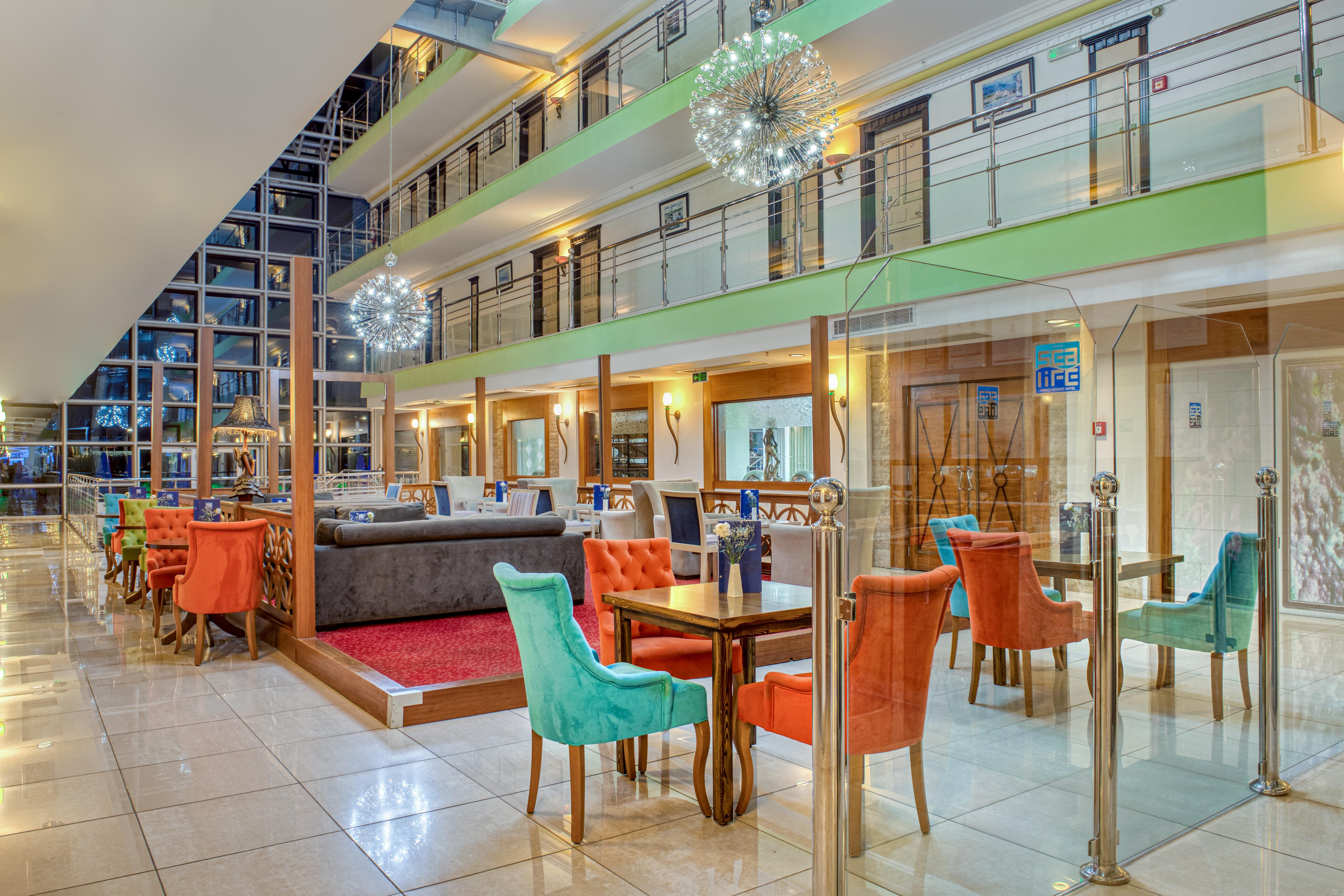 Sealife Family Resort Hotel Antalya Exterior photo The photo shows the interior of a modern hotel lobby or dining area. It features a spacious room with multiple levels visible. The decor includes colorful chairs in orange and turquoise, along with wooden tables. 

There are glass barriers separating